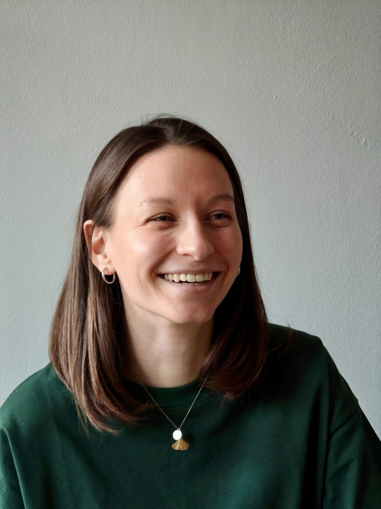 Pia steht vor einer grünen Wand. Man sieht ihr Porträt. Sie hat offene, braune Haare und lacht zur Seite.