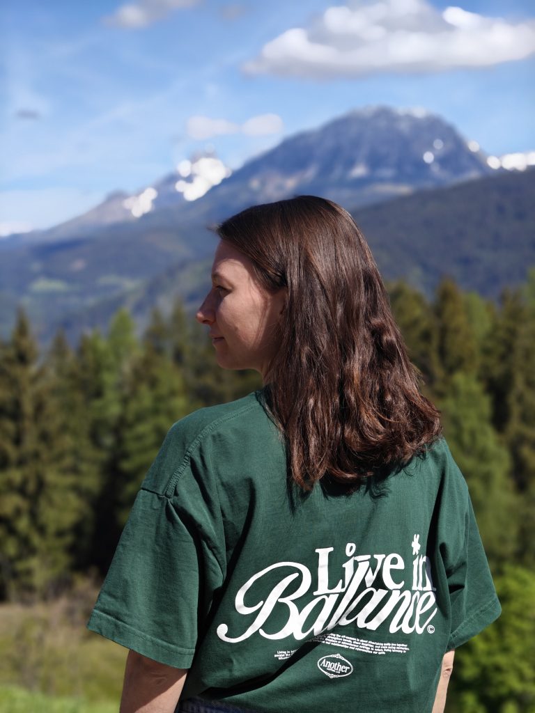 Eine Frage mit braunen Haaren und einem grünen Shirt steht vor einem Bergpanorame und schaut zur Seite nach links. Man sieht sie von hinten