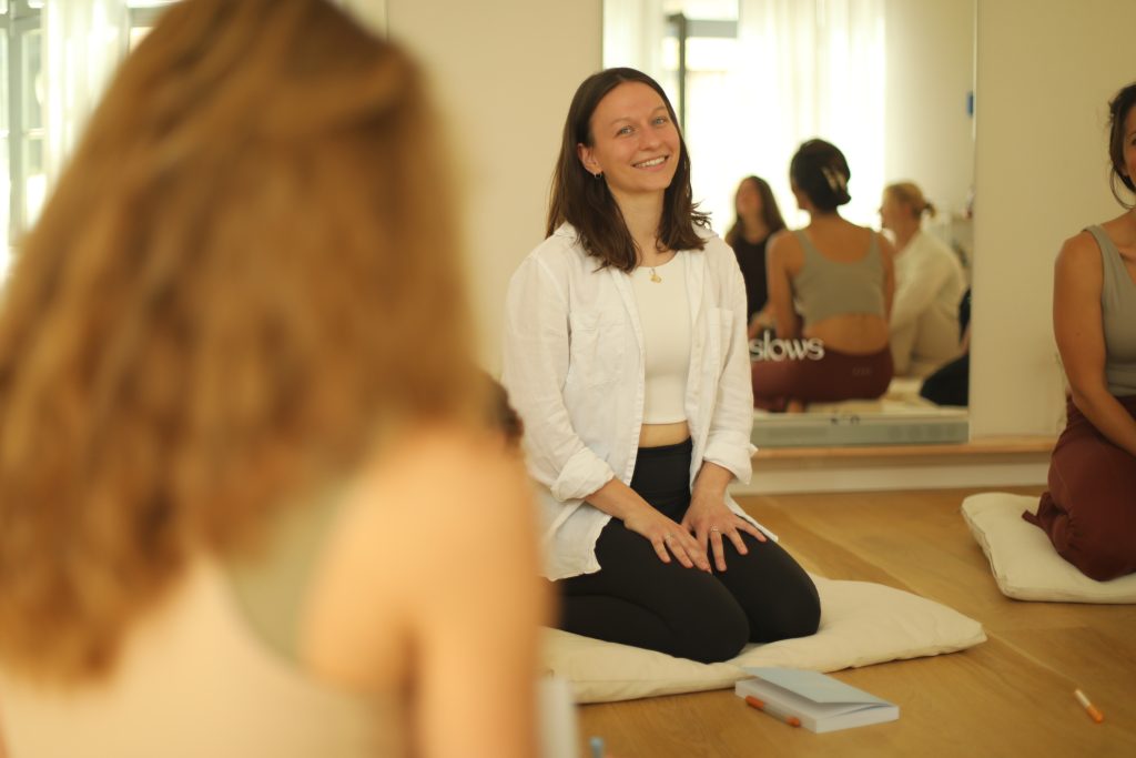Pia sitzt in einer weißen Bluse auf einer Meditationsmatte. Hinter ihr ist ein Spiegel, in dem man weiter sitzende Frauen sieht.