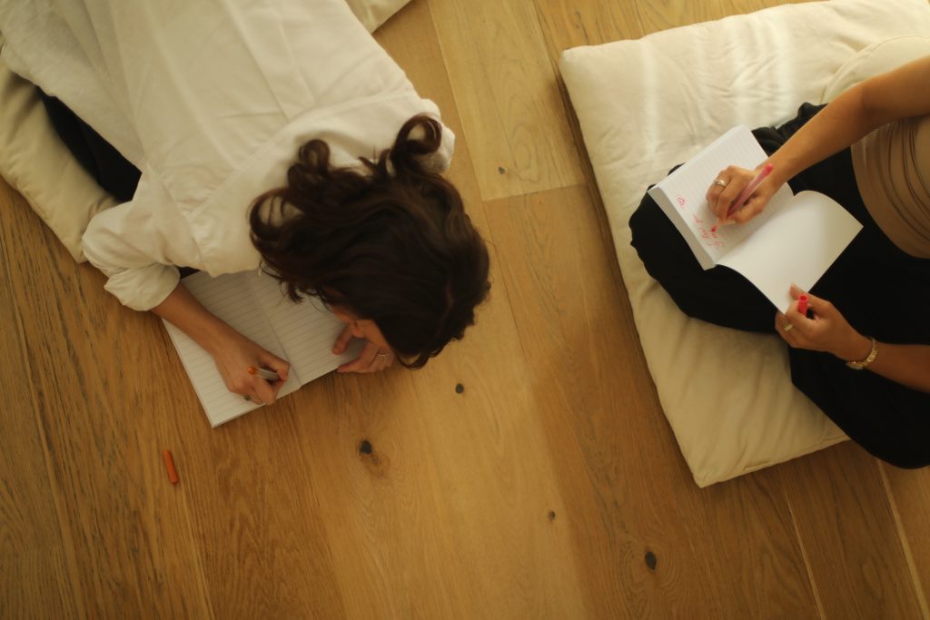 Zwei Frauen von oben fotografiert .Sie sitzen auf weißen Meditationsmatten auf einem Holzboden und schreiben beide jeweils in ein Notizbuch.