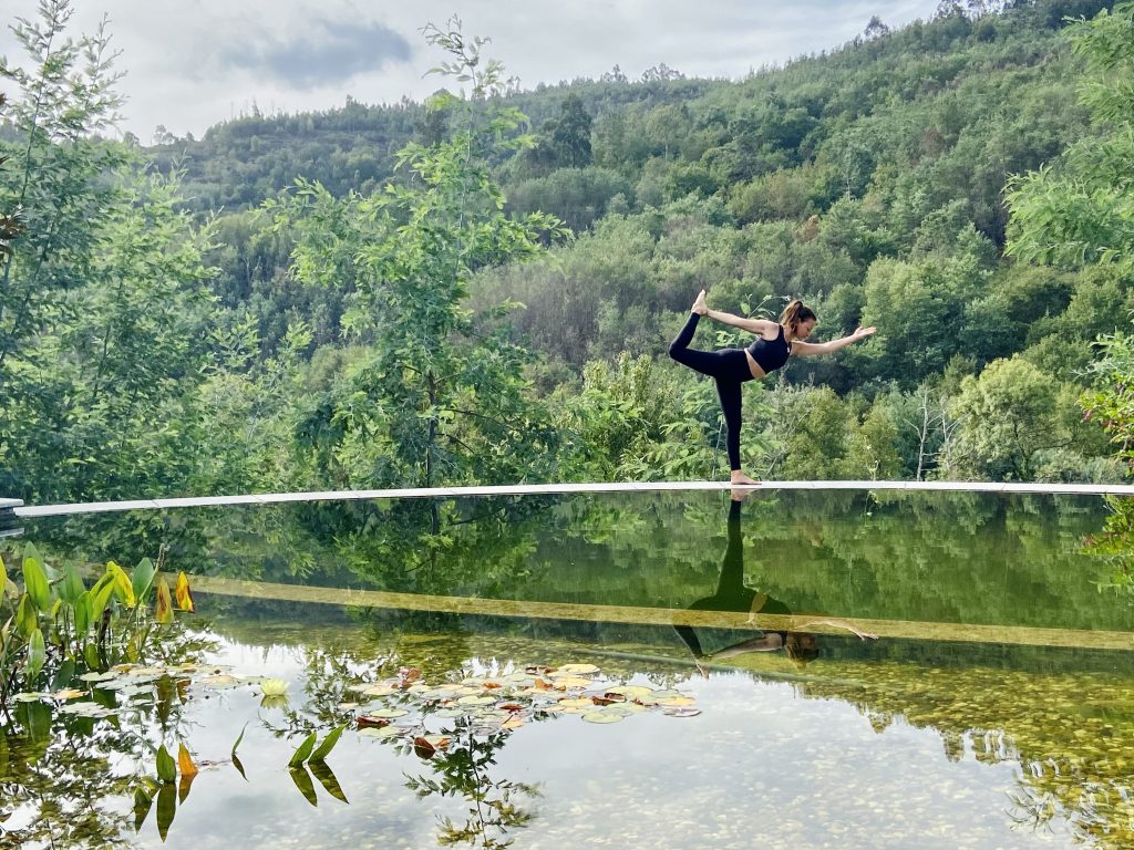 Pia steht am Ufer eines Teiches in der Pose des Yoga-Tänzers. Im Hintergrund ist ein Berg mit Wald zu sehen.