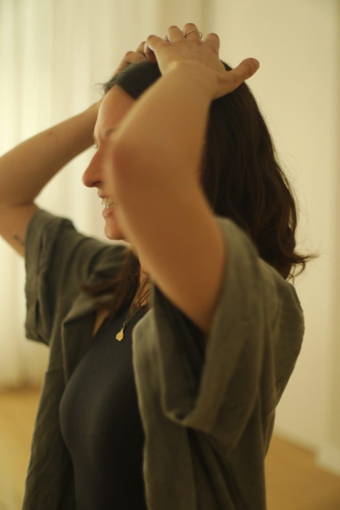 Pia sitz in einer grünen Jacke in einem Raum. Ihre Finger sind auf ihrem Kopf