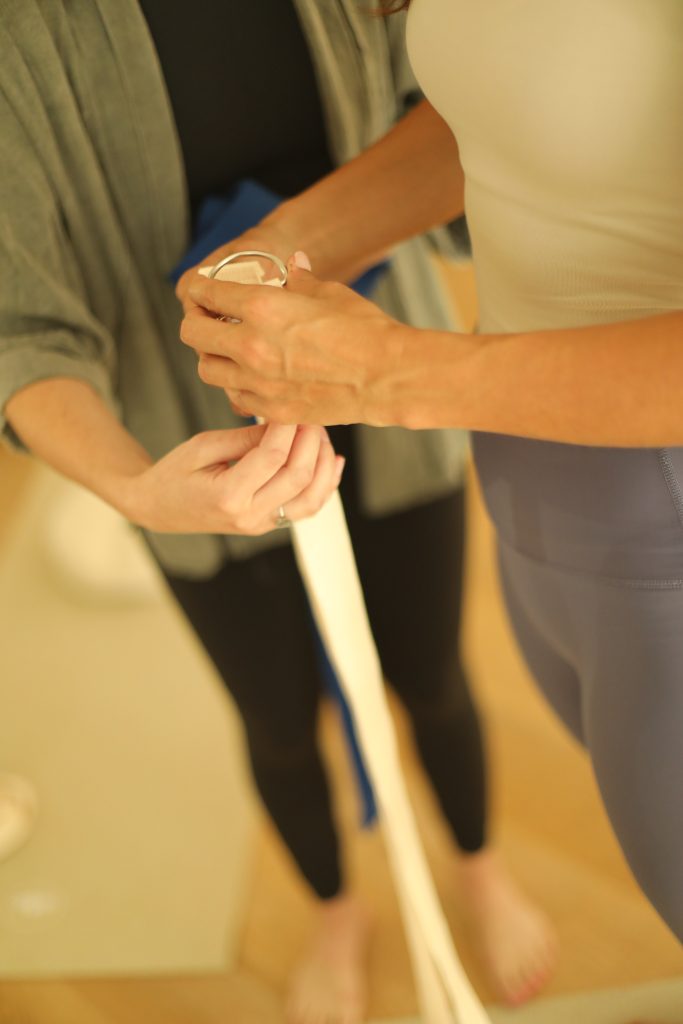 Man sieht zwei Frauen in Yoga Kleidung, die einen weißen Yogagurt halten. Ihre Gesichter sieht man nicht, nur ihre Körper.